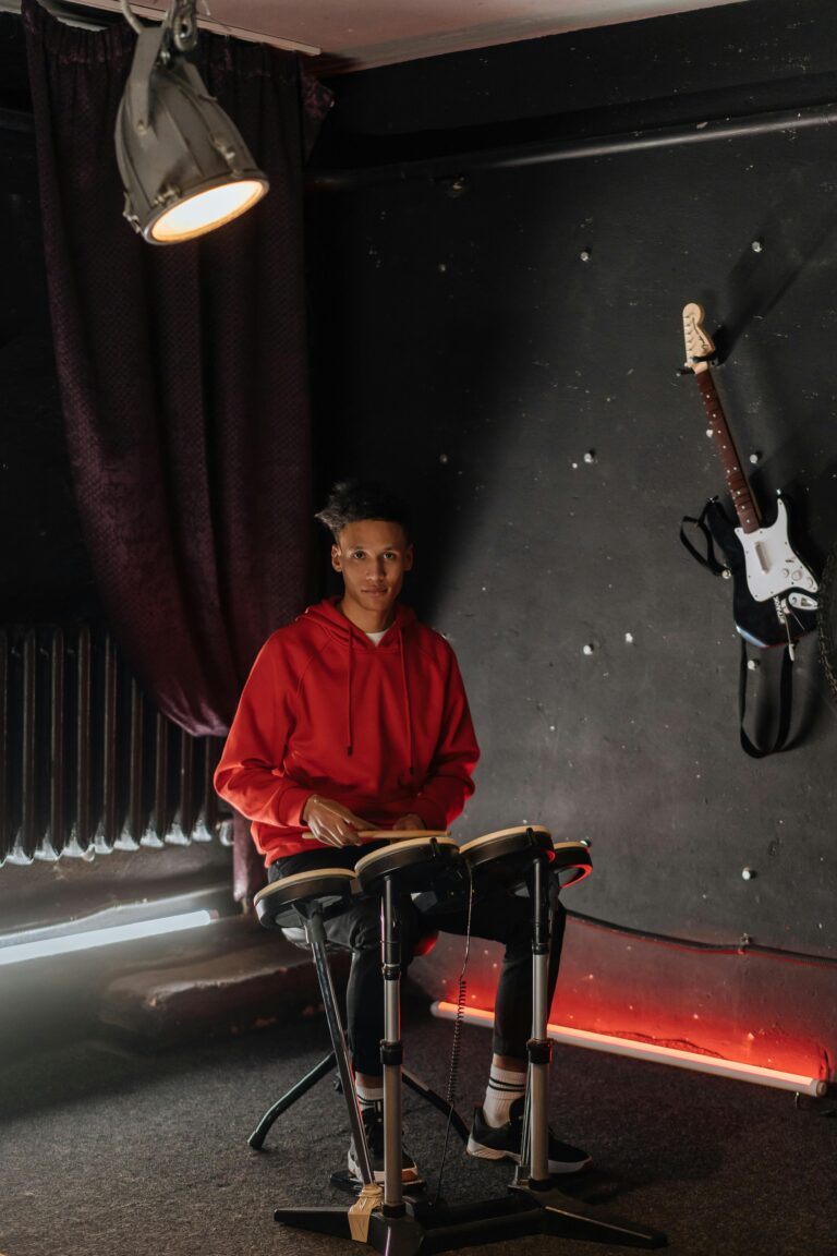 An electronic musician in a red hoodie playing drums indoors with a guitar on the wall.