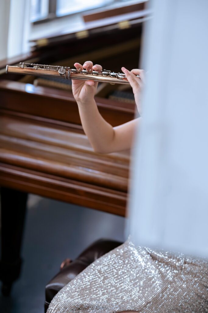 Crop female musician in elegant shiny dress sitting behind white curtain near piano and playing flute